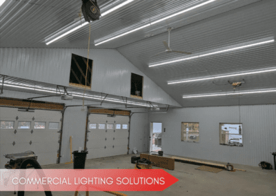 Large indoor workshop or garage space with multiple closed garage doors, efficient LED lighting on the ceiling, and a few scattered tools and materials on the floor. Text reads "Commercial Lighting Solutions.